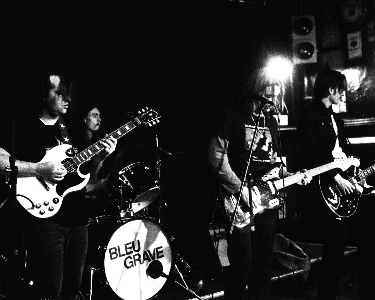 Bleu Grave on stage: two guitarists, the singer at the mic and the drummer behind a kick drum with the band name on it.