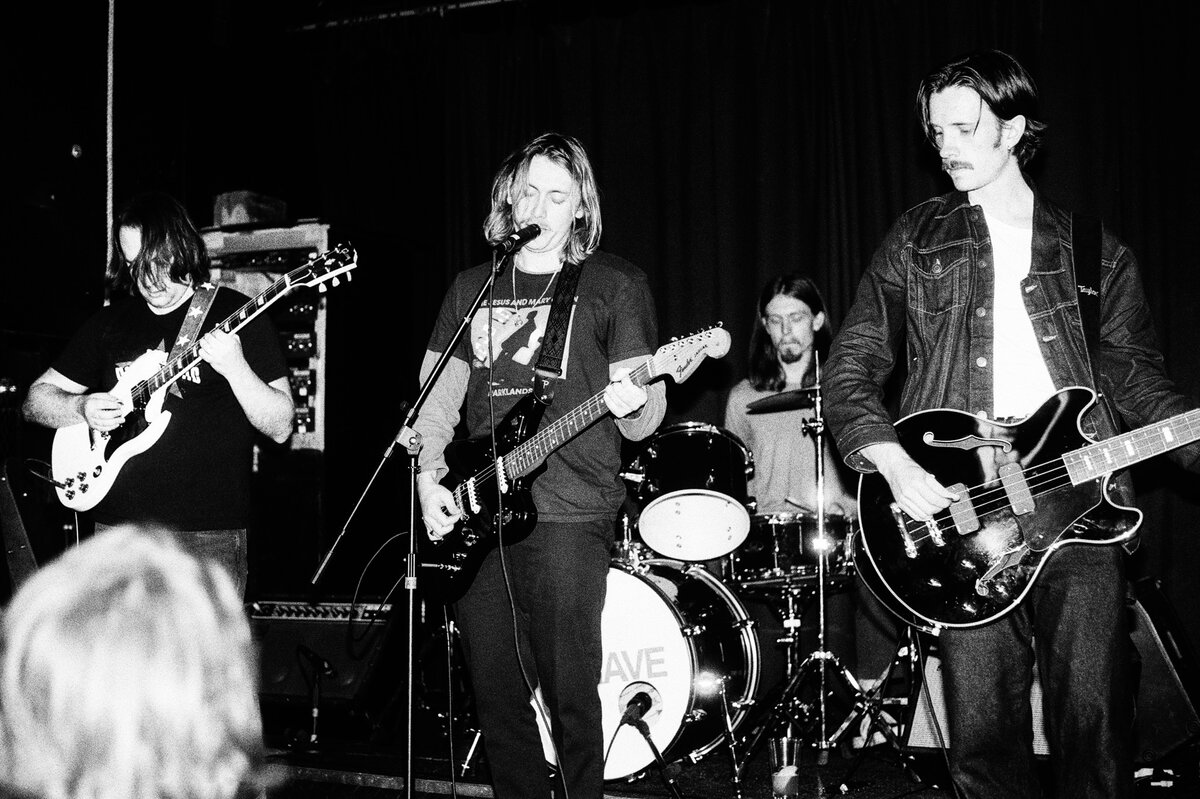 Bleu Grave playing a club show: guitarist on the left, the singer at the mic in the middle, the drummer behind them and the bassist in a denim jacket on the right.