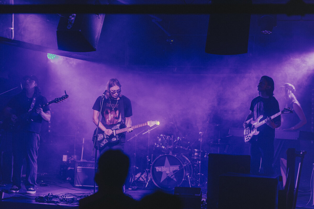 Bleu Grave across a smoke-filled stage: three guitarists spread out, the drum kit behind them with the band's star logo on the bass drum.